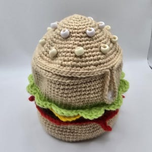 Op de afbeelding: Een gehaakte hamburger met een bruin broodje, groene sla, rode tomaat en gele kaas. De bovenste bol heeft witte versieringen. De burger is handgemaakt en lijkt een decoratief item te zijn.