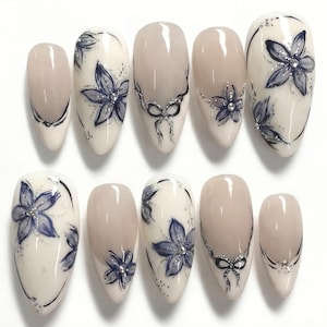 Pode incluir: Um conjunto de dez unhas postiças em formato amendoado com base nude brilhante. Os designs incluem flores azul-escuras pintadas à mão com detalhes em glitter prateado, pequenos strass transparentes e linhas pretas formando laços.