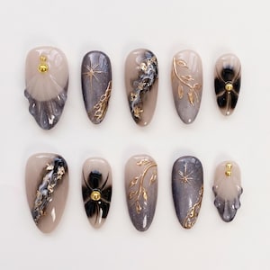 Peut inclure: Un ensemble de dix faux ongles aux designs variés. Les ongles présentent un mélange de tons neutres, beige, gris et noir, avec des accents dorés. Les motifs incluent des éclats d'étoiles, des motifs floraux et des volutes abstraites. Chaque ongle a une finition brillante.