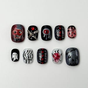 May include: A set of ten Halloween-themed press-on nails. Designs include a skull, a ghost, a dripping eye, and a heart, with accents of red, silver, and black. The nails are oval-shaped and have a glossy finish.