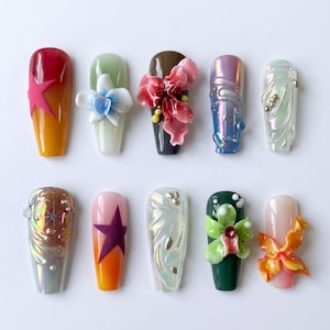 May include: Ten artificial nails with diverse designs are displayed. The nails showcase a variety of colours, such as red, orange, green, and pink, and are decorated with floral and star-shaped embellishments. Some nails have a glossy finish, while others have a pearlescent effect.