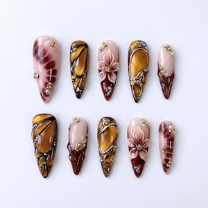 Può includere: Dieci unghie finte a forma di mandorla con disegni intricati. Le unghie presentano una varietà di colori, tra cui oro, marrone e rosa, con motivi floreali e astratti. Alcune unghie sono decorate con perle e dettagli dorati.