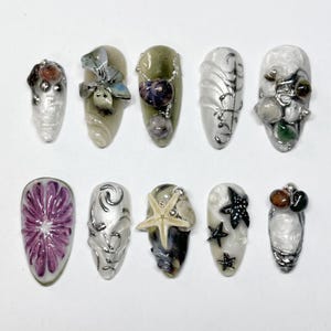 Könnte beinhalten: Zehn künstliche Fingernägel mit detaillierten Designs. Die Nägel weisen Verzierungen wie Muscheln, Seesterne und florale Akzente in Silber, Weiß, Grün und Lila auf. Die Nägel sind vor einem weißen Hintergrund angeordnet.