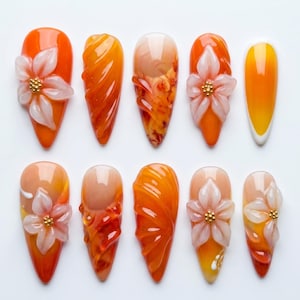 May include: A collection of ten artificial nails in various designs. The nails feature a gradient of orange, peach, and yellow hues. Some nails have 3D floral embellishments with gold accents, while others showcase textured patterns.