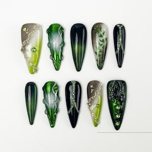 Puede incluir: Una colección de diez uñas artificiales en forma de almendra con varios diseños. Las uñas presentan tonos de verde, negro y plata, con adornos como cadenas, purpurina y patrones texturizados. Algunas uñas tienen un efecto degradado.