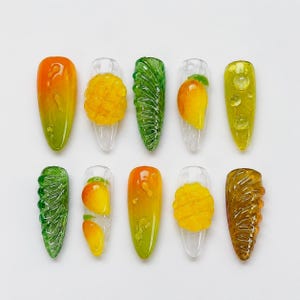 May include: Ten almond-shaped press-on nails featuring fruit-themed 3D designs. The set includes clear nails with raised yellow mango slices, green textured leaf patterns, and gradient orange-to-green bases with glossy water droplet accents.