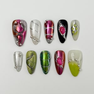 May include: A collection of ten almond-shaped artificial nails with various designs. The nails feature metallic silver, pink, green, and black bases, adorned with silver wire, beads, and abstract patterns. Some nails have a gradient effect.