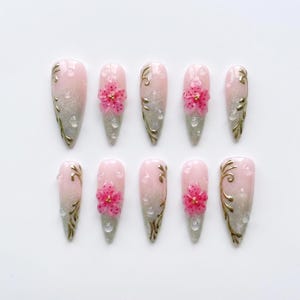 May include: A set of ten almond-shaped press-on nails featuring a light pink to silver glitter gradient base. Each nail is decorated with gold filigree swirls, clear raised water droplet accents, and some feature a three-dimensional pink flower.