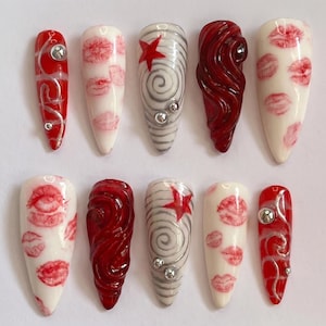 May include: A set of ten artificial nails with various designs. Some nails are red with silver swirls and rhinestones. Others are white with red lip prints, a red star, or a swirling gray design with rhinestones. One nail is a deep red, textured design.