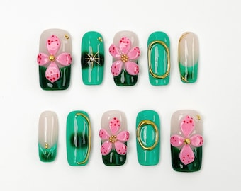 May include: A set of ten artificial nails with a variety of designs. Some nails feature a gradient of white to green, while others have a pink flower with red dots and gold accents. The nails are arranged on a white surface.