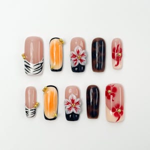 May include: A set of ten artificial nails in various designs. Some nails feature a nude base with black and white zebra stripes, while others have orange gradients, 3D flower accents, or dark brown and red patterns. Gold studs adorn some nails.