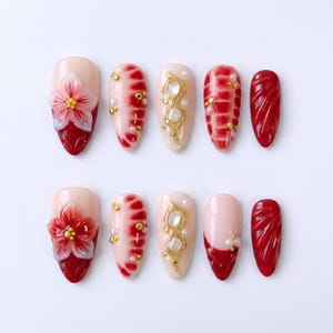 Può includere: Un set di dieci unghie finte con vari design. Alcune unghie presentano un fiore, altre hanno dettagli rossi e dorati, e alcune sono rosse piene. Le unghie sono ovali e hanno una finitura lucida.