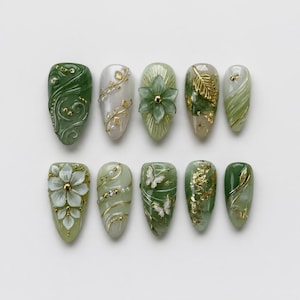 May include: A set of ten press-on nails in varying shades of sage and forest green, featuring intricate gold leaf accents, white floral embellishments, small pearls, and delicate butterfly motifs arranged in two horizontal rows on a white surface.