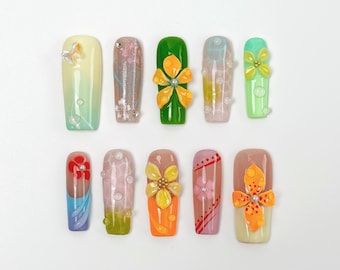 May include: A collection of ten artificial nails with various designs. The nails feature a range of colors, including green, pink, orange, and blue, and are adorned with floral and gem embellishments. Some nails have gradient effects.
