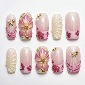 May include: A collection of ten artificial nails with diverse designs. The nails showcase pink, white, and gold hues. Some nails feature 3D floral and seashell decorations, while others have a French tip design with floral accents. The nails are displayed on a white surface.