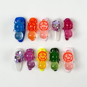 Puede incluir: Diez diseños de uñas con abalorios de tortugas translúcidas de colores. Los colores de esmalte de uñas incluyen azul, naranja, rosa, blanco, morado, amarillo y verde. Algunas uñas tienen adornos de cristal.