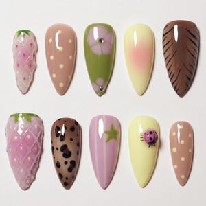 May include: A collection of ten artificial nails with various designs. The designs include strawberries, polka dots, floral patterns, animal prints, and a ladybug. The nails are in shades of pink, brown, green, and yellow.