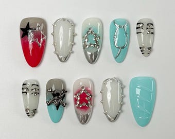 Puede incluir: Un conjunto de diez uñas artificiales con varios diseños. Las uñas presentan una mezcla de colores como rojo, gris, blanco y turquesa, con adornos como estrellas, calaveras y pedrería. Algunas uñas tienen detalles metálicos.