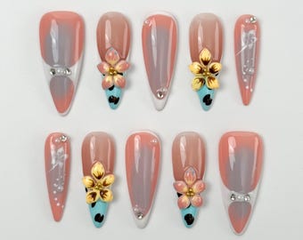 Op de afbeelding: Een set van tien kunstnagels in verschillende ontwerpen. De nagels hebben een verloop van perzik en grijs, met sommige versierd met 3D bloemaccenten in geel, roze en blauw. Sommige nagels hebben kleine kristallen versieringen.