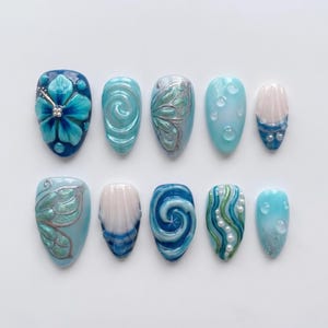 Könnte beinhalten: Zehn künstliche Fingernägel mit verschiedenen Designs. Die Nägel zeigen Blau-, Weiß- und Silbertöne mit Blumen-, Wirbel- und Schmetterlingsmustern. Einige Nägel haben 3D-Verzierungen, darunter Perlen und Wassertropfeneffekte.