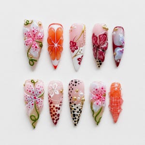 Op de afbeelding: Een set van tien kunstnagels met spitse punten, voorzien van diverse ontwerpen waaronder roze en witte bloemmotieven, oranje citrus-geïnspireerde segmenten, gouden accenten, witte parels en zwarte stippen op een lichtroze basis.