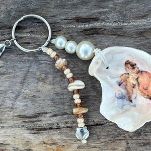 Puede incluir: Un llavero con anillo y mosquetón de metal plateado, una hilera de pequeñas cuentas marrones y crema que termina en un dije de concha plateada, tres perlas blancas y una concha de ostra pintada con una pequeña tortuga marina naranja.