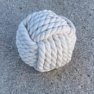 May include: A white rope knot, tied into a ball shape. The knot is made of thick, white rope and is a decorative item.