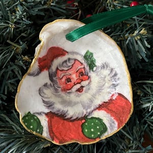 Puede incluir: Adorno navideño con forma de concha de ostra, con una ilustración vintage de Papá Noel. El borde es dorado y tiene una cinta verde para colgarlo. La imagen de Papá Noel es roja, blanca y verde.