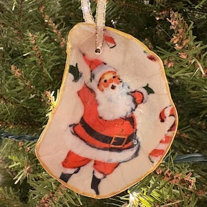 Pode incluir: Um enfeite de Natal em forma de concha, com uma ilustração vintage do Papai Noel em traje vermelho e barba branca. O enfeite tem uma borda dourada e uma fita para pendurar.