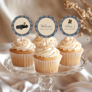 Könnte beinhalten: Drei Cupcakes mit runden Papieraufsteckern, die einen blauen Karorand und eine beige Mitte haben. Die Aufstecker zeigen einen Teddybären im Auto, den Text Welcome BABY WILLIAMS und den Text Olivia's BABY SHOWER mit einer Pferdeabbildung.