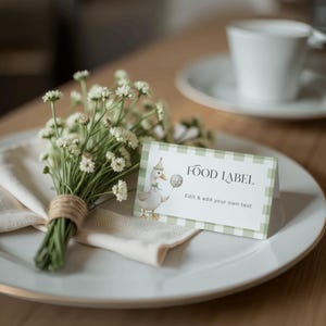 May include: A white plate with a gold rim holds a folded beige napkin, a small bouquet of white flowers tied with twine, and a green and white checkered food label. The label features a cartoon goose.