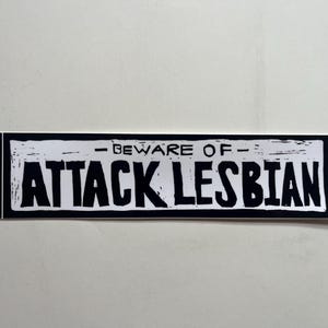 May include: A black and white sticker with the text "- BEWARE OF - ATTACK LESBIAN" in bold, distressed lettering. The sticker has a rectangular shape with a black border and a white background.