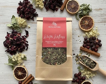 Könnte beinhalten: Ein Kraftpapierbeutel mit "Winter Wellness" Tee mit rotem Etikett, umgeben von getrockneten Orangenscheiben, Zimtstangen und getrockneten Blüten. Die Teemischung enthält Hibiskus und Orangenschalen. Auf einer weißen Holzoberfläche angeordnet.