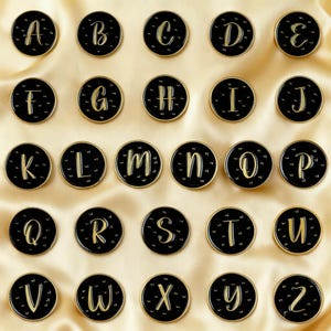 Może przedstawiać: Kolekcja czarnych i złotych przypinek z literami alfabetu ułożonych na złotej satynowej powierzchni. Każda przypinka przedstawia inną literę alfabetu w złotym kroju pisma, otoczoną złotą obwódką. Przypinki są okrągłe i mają błyszczące wykończenie.
