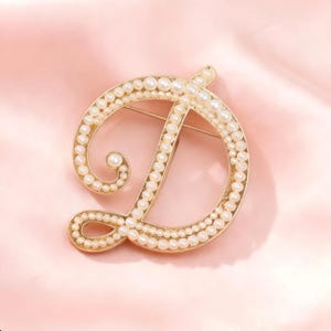 Può includere: Una spilla in metallo color oro a forma di lettera maiuscola D, caratterizzata da un bordo decorativo incastonato con piccole perle bianche rotonde. La lettera è disegnata in uno stile corsivo con un'evidente asola nella parte superiore sinistra.
