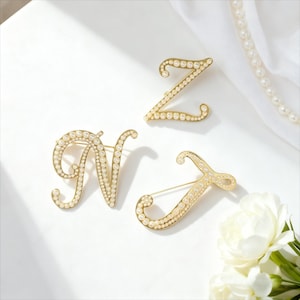 May include: Three gold-toned letter brooches: "Z", "N", and "J", each encrusted with small, round, off-white pearls. The brooches are arranged on a white surface, with a pearl necklace and white flowers in the background.
