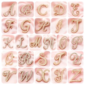 Może przedstawiać: Kolekcja broszek z inicjałami w odcieniach złota, każda litera alfabetu od A do Z, inkrustowana małymi, okrągłymi, białymi perłami. Broszki są prezentowane na różowym satynowym tle.