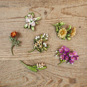 May include: Six floral brooches in different designs are displayed. The brooches are crafted with detailed enamel work in shades of pink, orange, yellow, and purple, with green accents and faux pearls. Each brooch represents a unique floral arrangement.