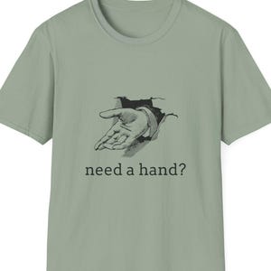 May include: Sage green t-shirt featuring a black and white illustration of a hand reaching out from a torn surface. The text "need a hand?" is printed below the graphic. A casual, everyday wear tee.