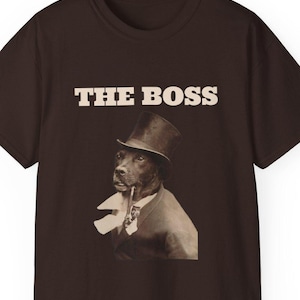 May include: Brown t-shirt featuring a sepia-toned image of a dog in a top hat and suit, smoking a pipe. The text "THE BOSS" is printed above the dog's head in a bold, sans-serif font.