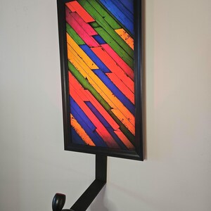 May include: A framed artwork with a vibrant, multicolored abstract design. The artwork features diagonal wooden planks in shades of red, orange, yellow, green, and blue. A black metal bracket supports the frame, with a black control knob attached.