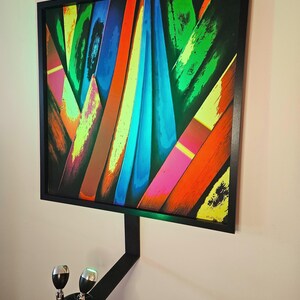 May include: Abstract art print in a black frame, featuring a vibrant composition of colorful, overlapping shapes. The artwork includes shades of green, blue, orange, and pink. A black metal bracket with two lights is mounted below the artwork.