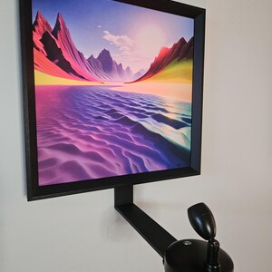 May include: A framed landscape art print featuring a colorful desert scene with mountains and a body of water. The artwork is in a black frame and mounted on a wall with a black adjustable spotlight.