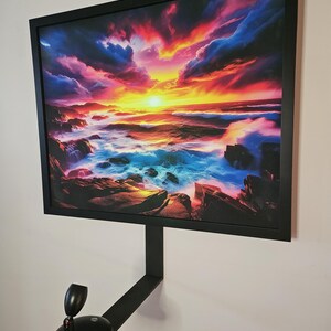 May include: A framed art print depicting a vibrant sunset over a rocky coastline. The artwork features a dramatic sky with shades of red, orange, and blue, reflecting on the ocean waves. The frame and mounting hardware are black.