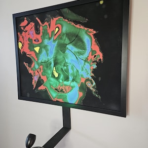 May include: Abstract art piece in a black frame, featuring a vibrant mix of colors including green, blue, red, and yellow against a black background. The artwork is mounted on a wall with a black, adjustable spotlight.