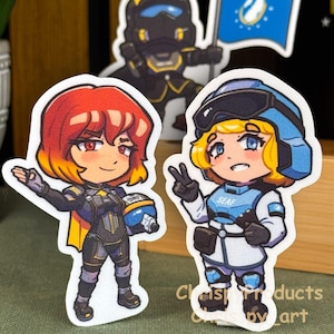 May include: Three die-cut stickers featuring chibi-style characters in tactical gear. One character has red hair and black armor, another has blonde hair and a blue and white uniform with SEAF text, and a third holds a blue flag with a white emblem.