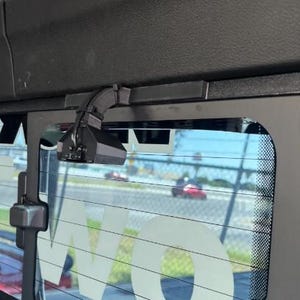 May include: A black, angular, 3D-printed mounting bracket attached to the top frame of a sliding truck rear window. The mount features a curved arm extending downward to hold a small, dark, multi-faceted electronic device with visible wiring.