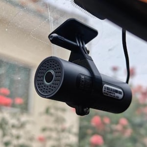 Può includere: Una dash cam cilindrica nera montata sul parabrezza di un veicolo. Il dispositivo presenta un obiettivo circolare circondato da una griglia per altoparlante perforata sulla parte anteriore, una staffa di montaggio nera e un'etichetta bianca.