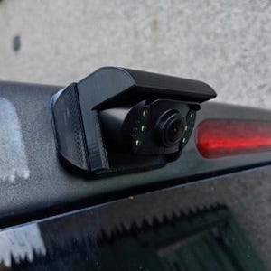 May include: A black 3D-printed mounting bracket attached to a vehicle, holding a small black backup camera with a circular lens and several small LED lights surrounding the lens, positioned next to a red horizontal tail light on the vehicle body.