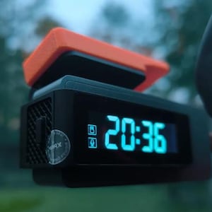 May include: A black digital dash cam featuring a bright cyan LED display showing the time 20:36, with two small icons for memory and microphone. The device has a textured side panel and a prominent orange plastic top component attached to the unit.